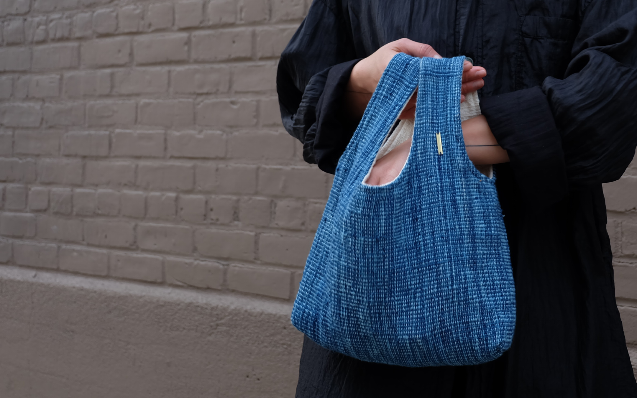 A person wearing a black textured shirt holds a blue woven tote bag with double handles, standing against a gray brick wall.